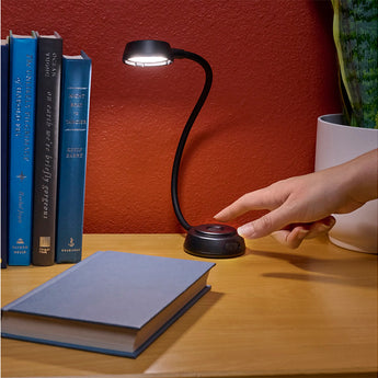 Hand adjusting a flexible desk lamp on a table with books and a plant in the background.